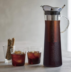 Tall glass cold brew pitcher filled with dark coffee, accompanied by two glasses of iced cold brew and coffee beans