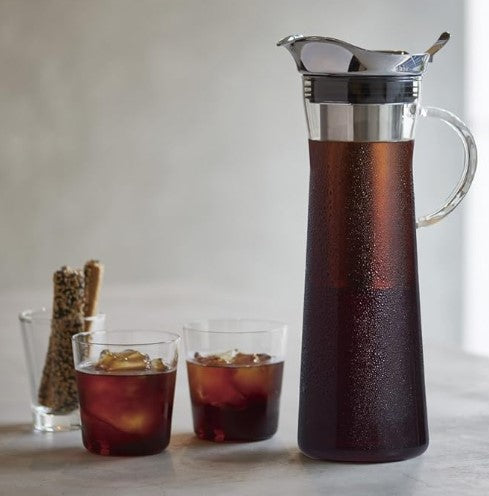Tall glass cold brew pitcher filled with dark coffee, accompanied by two glasses of iced cold brew and coffee beans