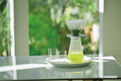 Cha Cha Tea Dripper with glass cone sitting on white plate with green tea brewing, two empty glasses beside it, outdoor