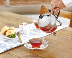 Person pouring red tea from glass Chaor teapot into matching cup at wooden table with pastries.
