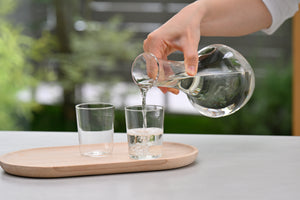 Pouring clear liquid from glass carafe into drinking glass on wooden tray outdoors.