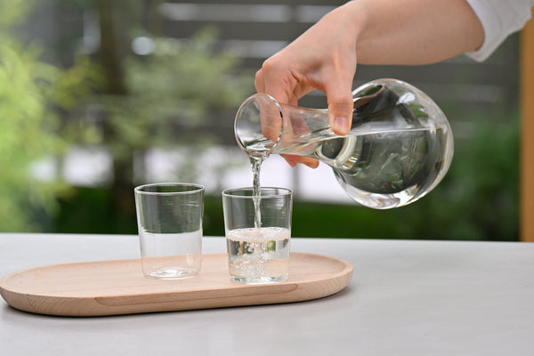 Pouring clear liquid from glass carafe into drinking glass on wooden tray outdoors.
