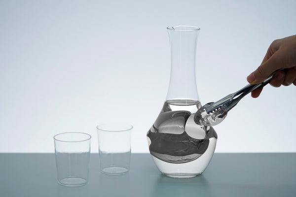 Hand pouring dark liquid into glass carafe with ice well, flanked by two empty drinking glasses.