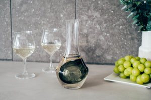 Glass carafe with dark liquid in ice well, displayed with two wine glasses and green grapes.