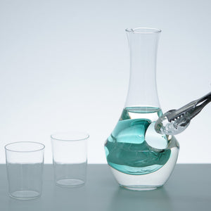 Hand pouring turquoise liquid into glass carafe with ice well, next to two empty drinking glasses.