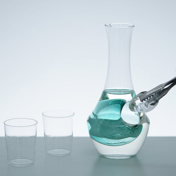 Hand pouring turquoise liquid into glass carafe with ice well, next to two empty drinking glasses.