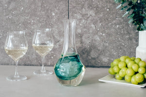 Glass carafe with turquoise liquid in ice well, styled with wine glasses and fresh green grapes.