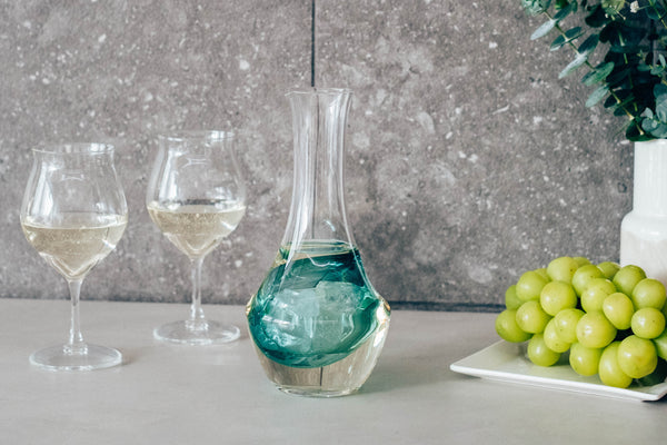 Glass carafe with turquoise liquid in ice well, styled with wine glasses and fresh green grapes.