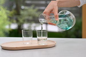 Pouring turquoise-tinted liquid from glass carafe into drinking glass on wooden tray.