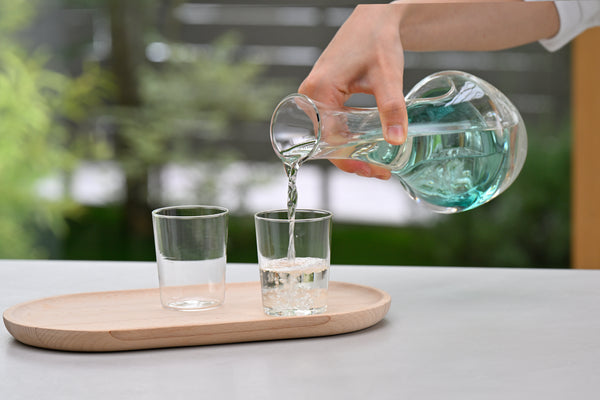 Pouring turquoise-tinted liquid from glass carafe into drinking glass on wooden tray.