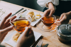 Two people toasting with amber tea in glass cups alongside pastries and cheese on wooden table.