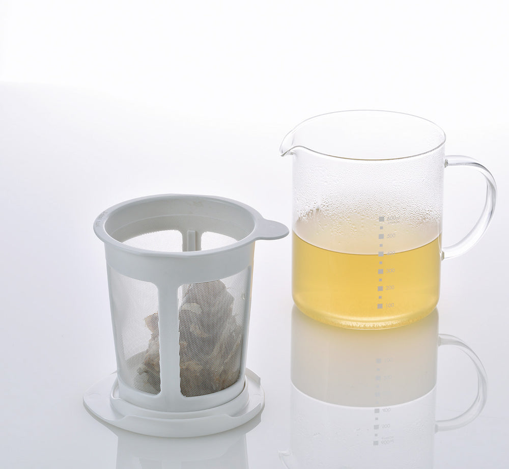 Glass pitcher filled with pale yellow broth next to removable mesh strainer basket containing tea bag.