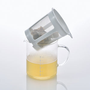 Mesh strainer basket tilted over glass pitcher of broth, demonstrating how to hook strainer on rim.