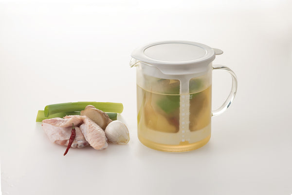 Glass pitcher with lid containing golden broth and steeped ingredients, displayed with fresh ginger, scallion, and chili.