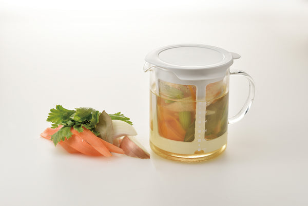 Glass pitcher with lid containing light broth and steeped vegetables, shown alongside fresh carrot and parsley.