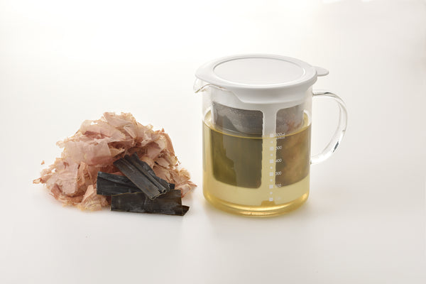Glass pitcher with lid containing darker broth and mesh strainer, paired with dried seafood ingredients on white surface.