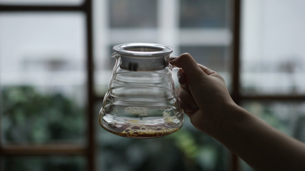 Person pouring dark coffee from glass V60 server into clear mug with ice, surrounded by fresh berries and pastries on table.