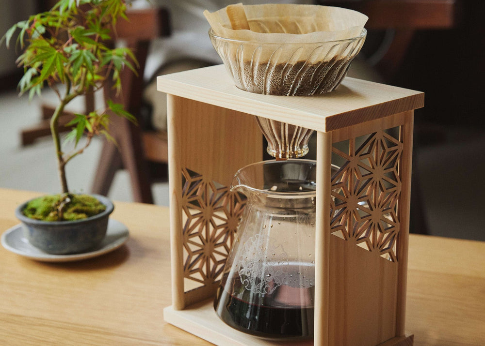 Glass coffee dripper with glass carafe on a wooden stand, with a plant in the background.