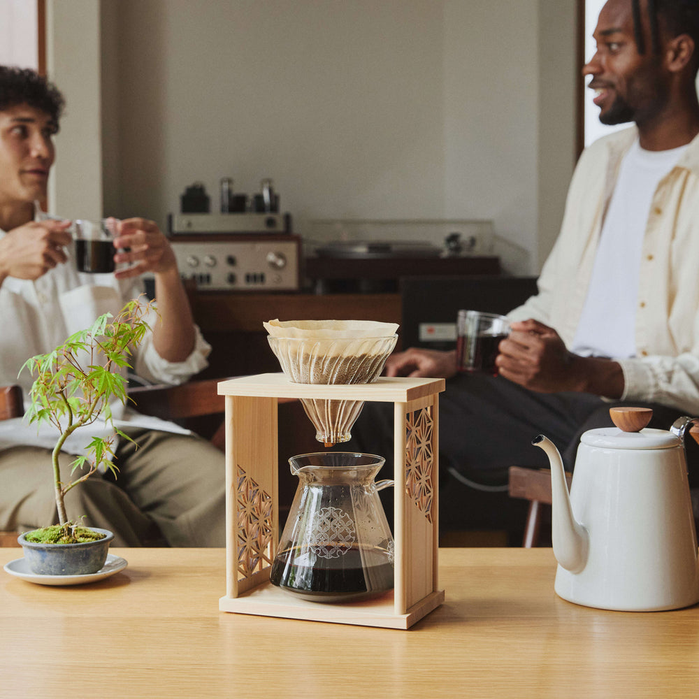 Glass coffee dripper with glass carafe on a wooden stand, with a plant in the background.
