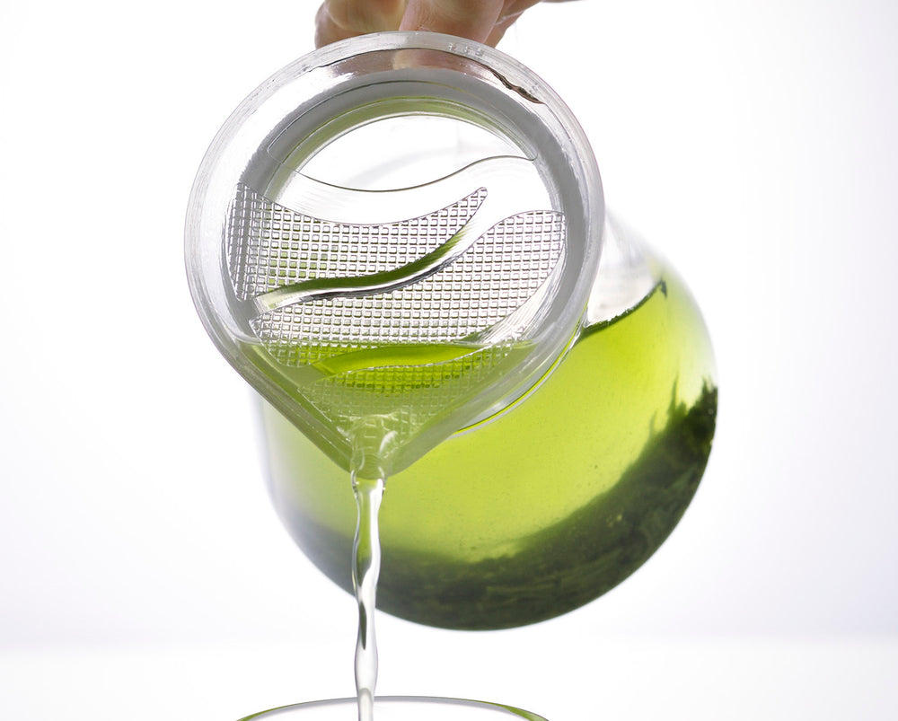Glass teapot with brewed green tea and a matching glass cup on outdoor table with foliage background.