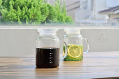 Two glass jugs on wooden windowsill: one filled with dark coffee or tea, another with lemon-infused water