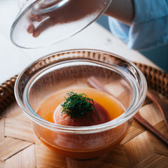 Clear glass rice bowl filled with amber broth and topped with scallop and green seaweed garnish on wooden surface.