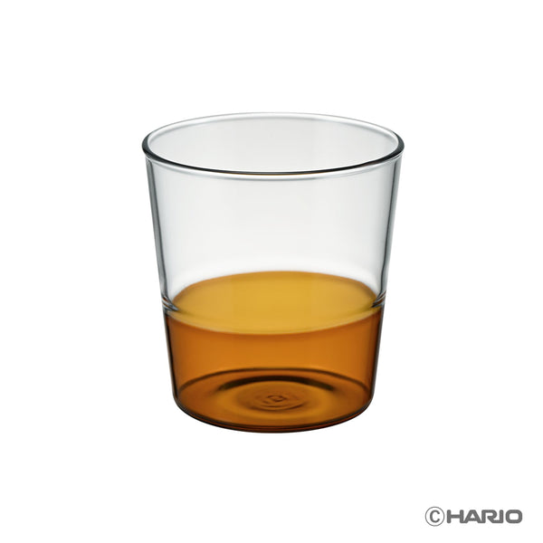Clear HARIO drinking glass half-filled with amber-colored liquid against white background.