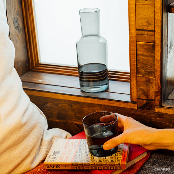 Person holding dark liquid in glass cup while carafe sits on wooden window sill in background.