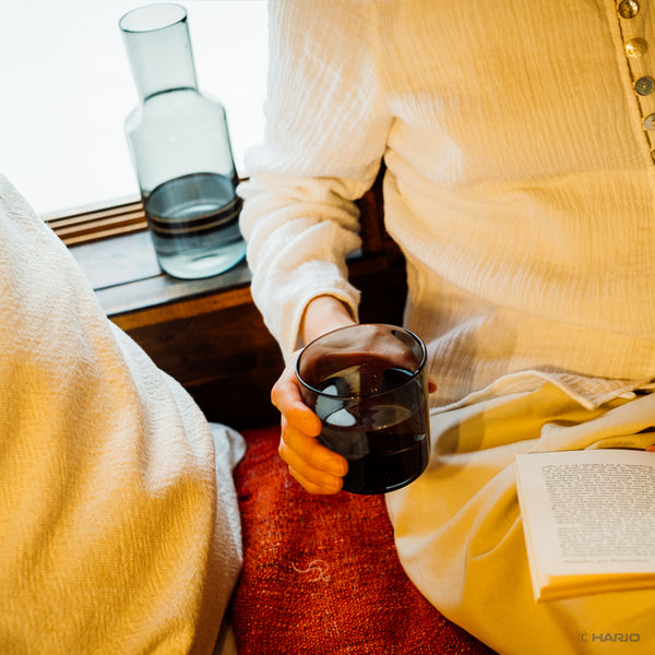 Person in yellow sweater holding dark-filled glass cup with carafe visible on window sill.