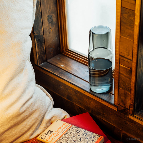 Wooden window sill with clear glass carafe filled with water and matching glass cup beside a red book.