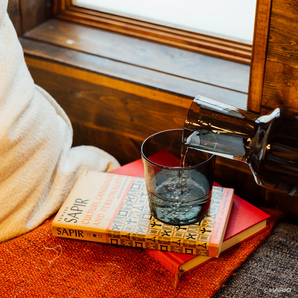 Clear liquid being poured from dark glass carafe into cup on top of stacked books.