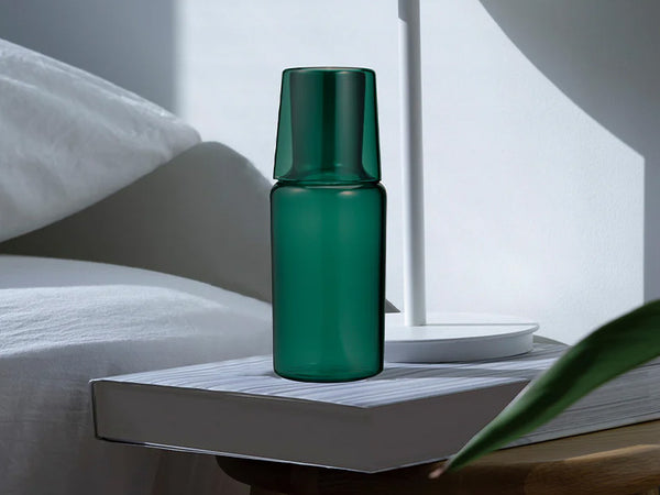 Dark green glass carafe with matching cup standing upright on a book in bright sunlight.