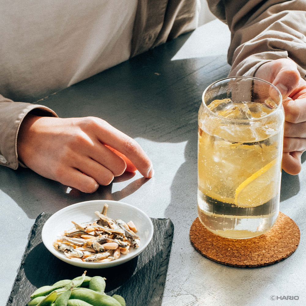Two people clinking glasses of yellow citrus drinks with ice and lemon slices, with edamame and small fish appetizers