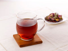 Clear glass mug filled with deep red tea on wooden coaster, dried flowers in background.