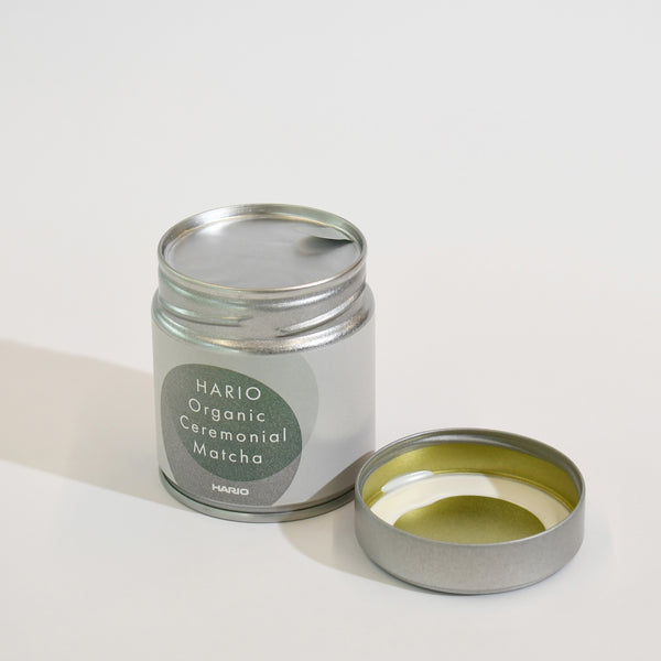 Opened tin with matcha powder inside and silver measuring scoop displayed separately.