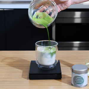 Hand pouring prepared matcha from glass bowl into cup of ice water.