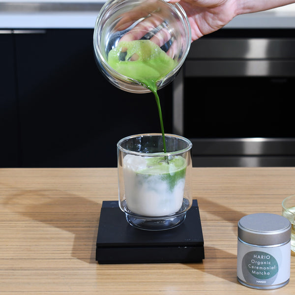 Hand pouring prepared matcha from glass bowl into cup of ice water.