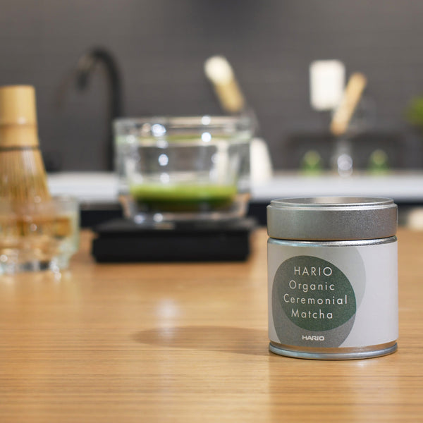 HARIO Organic Ceremonial Matcha tin positioned on wooden counter near matcha whisk.