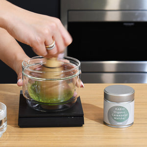 Hand whisking matcha in glass bowl on heated base with bamboo whisk.