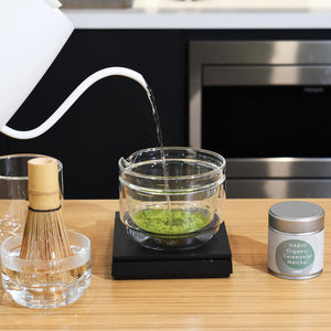 Hot water being poured into glass bowl containing matcha powder.
