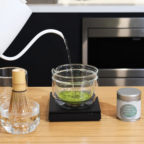 Hot water being poured into glass bowl containing matcha powder.