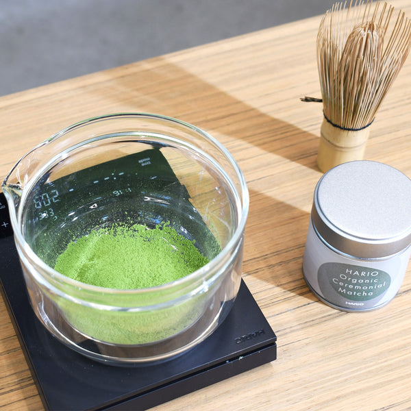 Heated glass bowl filled with matcha powder next to tin and bamboo whisk.