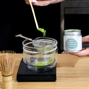 Bamboo whisk lifting frothy matcha from bowl while tin sits nearby.