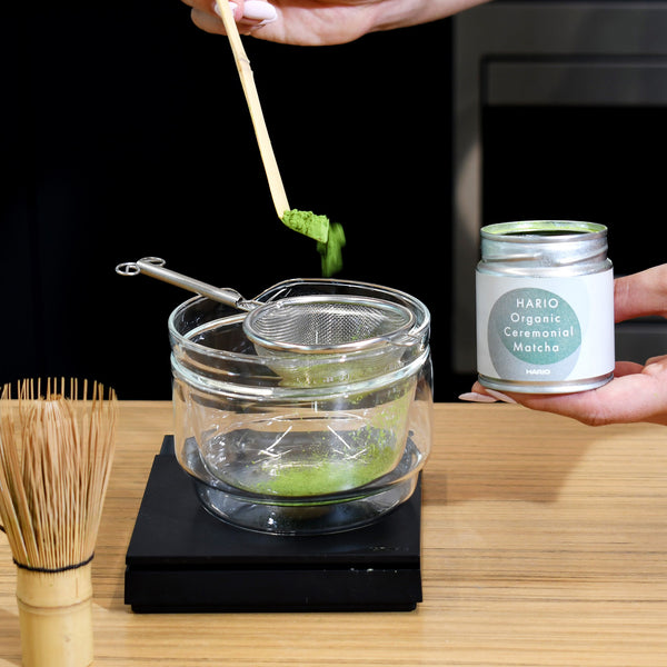 Bamboo whisk lifting frothy matcha from bowl while tin sits nearby.