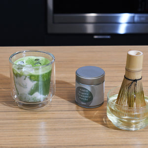 Completed matcha drink with foam in glass, tin, and bamboo whisk displayed together.
