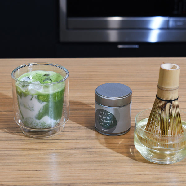 Completed matcha drink with foam in glass, tin, and bamboo whisk displayed together.