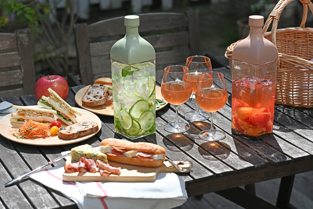Outdoor table set with cucumber and herbal infused Ka-Ku bottle alongside wine glasses and appetizers.