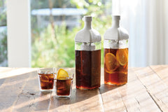 Two tall white bottles with dark cold brew coffee and two glasses with ice and lemon slices on wooden table near window.