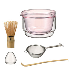 Pink-tinted glass matcha bowl with matching accessories including bamboo whisk, metal sieve, small glass cup, and wooden