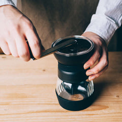 Person's hands holding black ceramic Skerton Pro grinder with glass lower chamber on wooden table